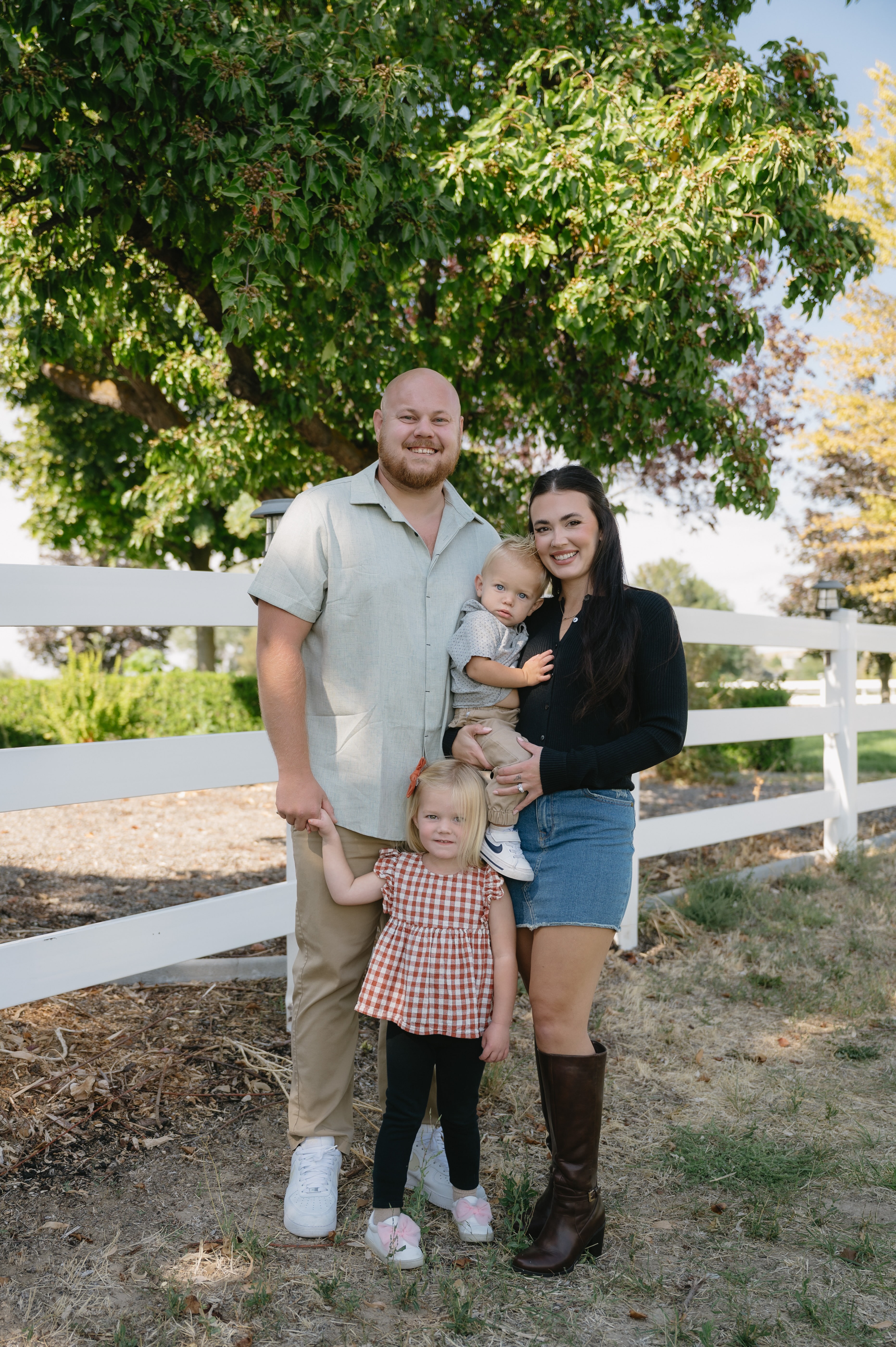 Smiling family of four with father, mother holding toddler, and young girl standing outdoors by a white fence and green trees.