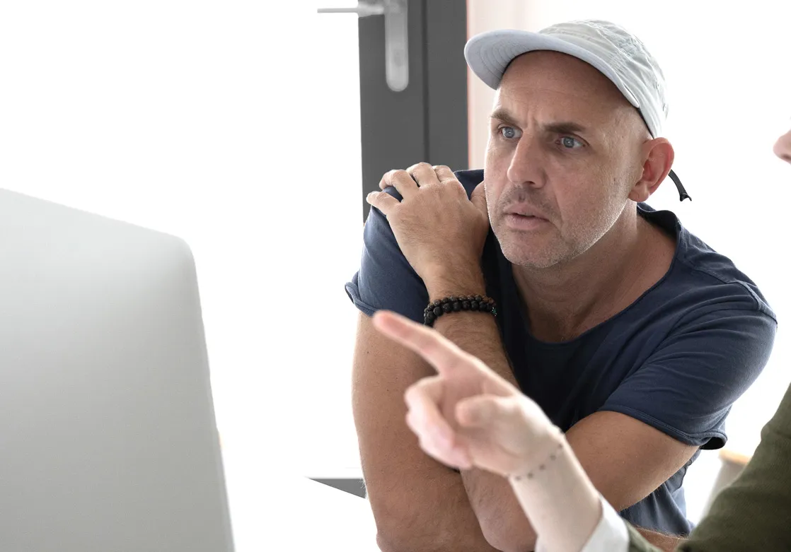 Ein Mann mit Kappe schaut aufmerksam auf einen Bildschirm, während eine andere Person auf den Bildschirm zeigt.