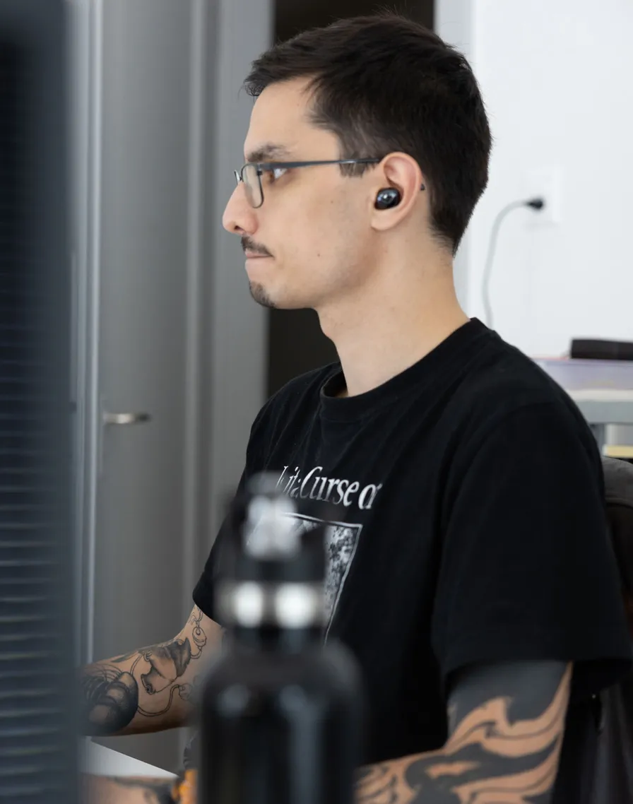 Seitliches Porträt eines Mannes mit Brille, Ohrhörern und tätowierten Armen, der konzentriert am Computer arbeitet.