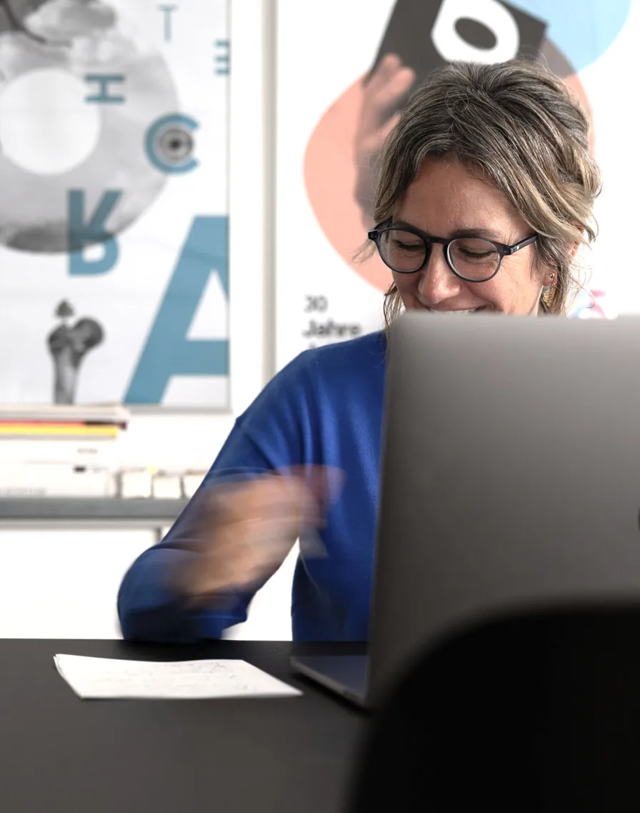 Frau mit Brille und blauem Pullover lächelt vor einem Laptop in einem modernen Büro.