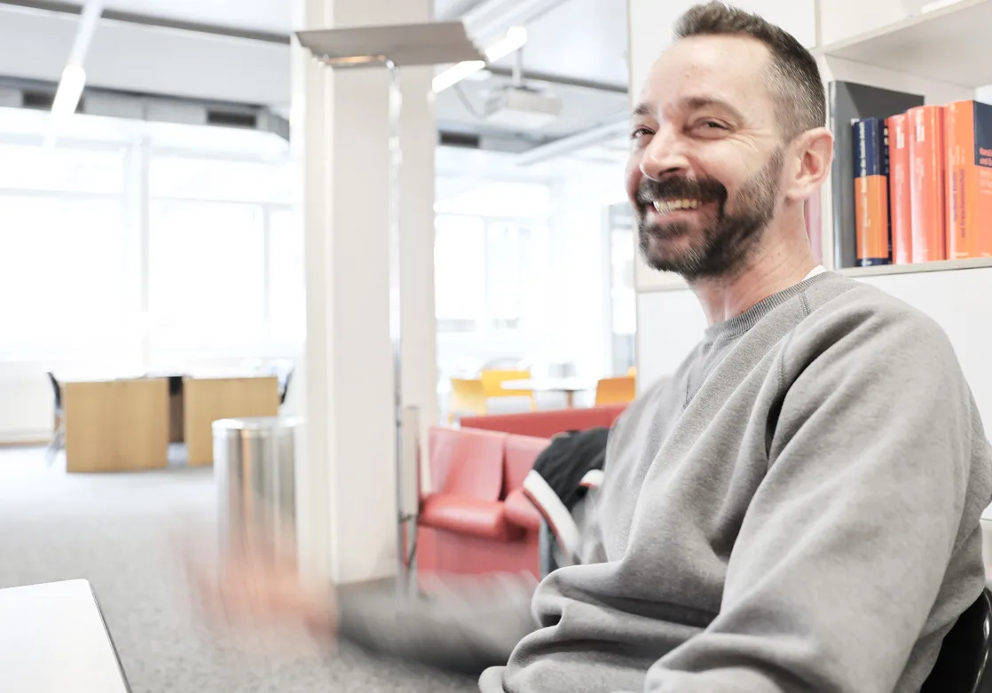 Lächelnder Mann mit Bart und grauem Sweatshirt in einem hellen Büro mit Bücheregal und roten Sofas.