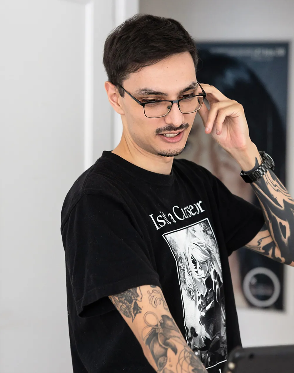 Damin Samsoodeen mit Brille und Tätowierungen trägt ein schwarzes T-Shirt mit Grafik und schaut beim Berühren seiner Brille nach unten.