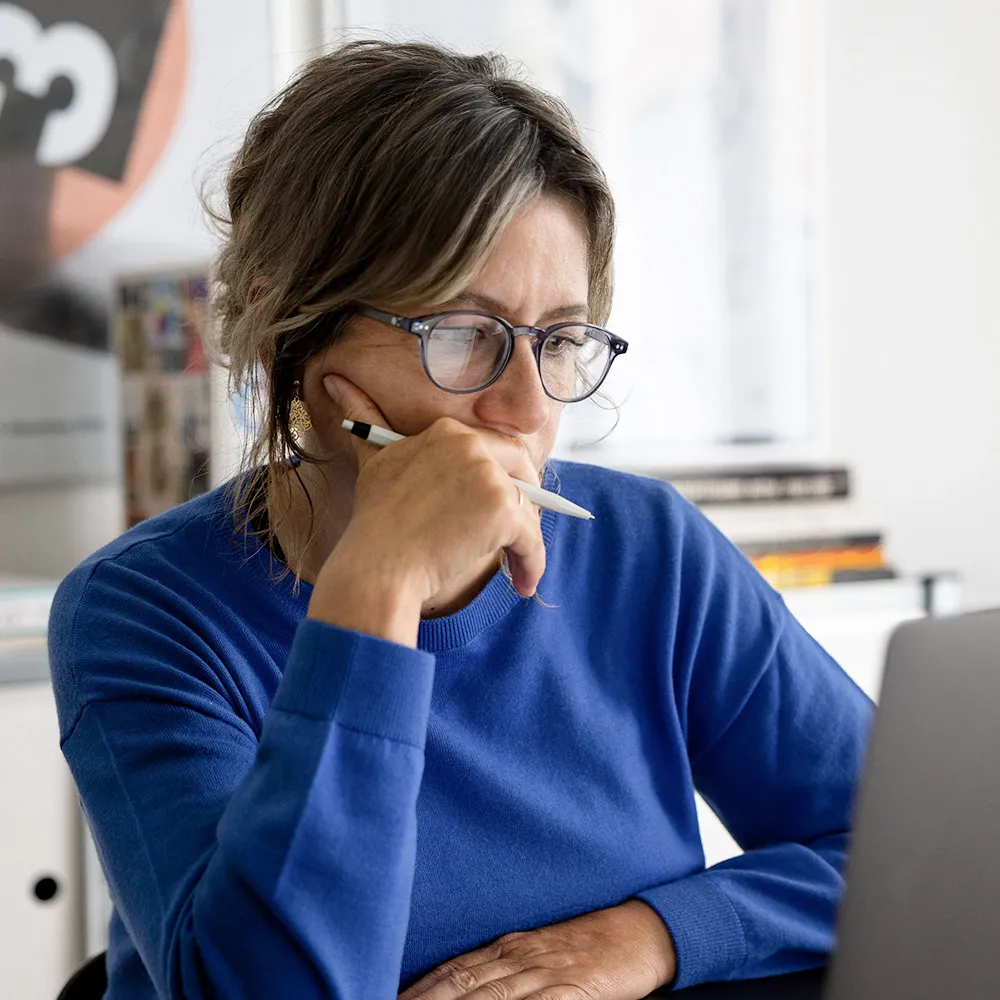 Sandra Mazenauer mit Brille und blauem Pullover konzentriert sich auf einen Laptop und hält einen Stift in der Hand.