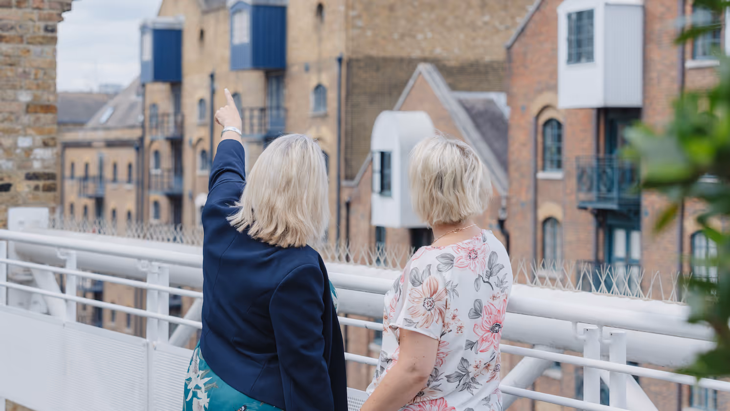 Two women are standing on a rooftop. One is pointing towards something in the distance. The other is looking at the object she is pointing towards. 