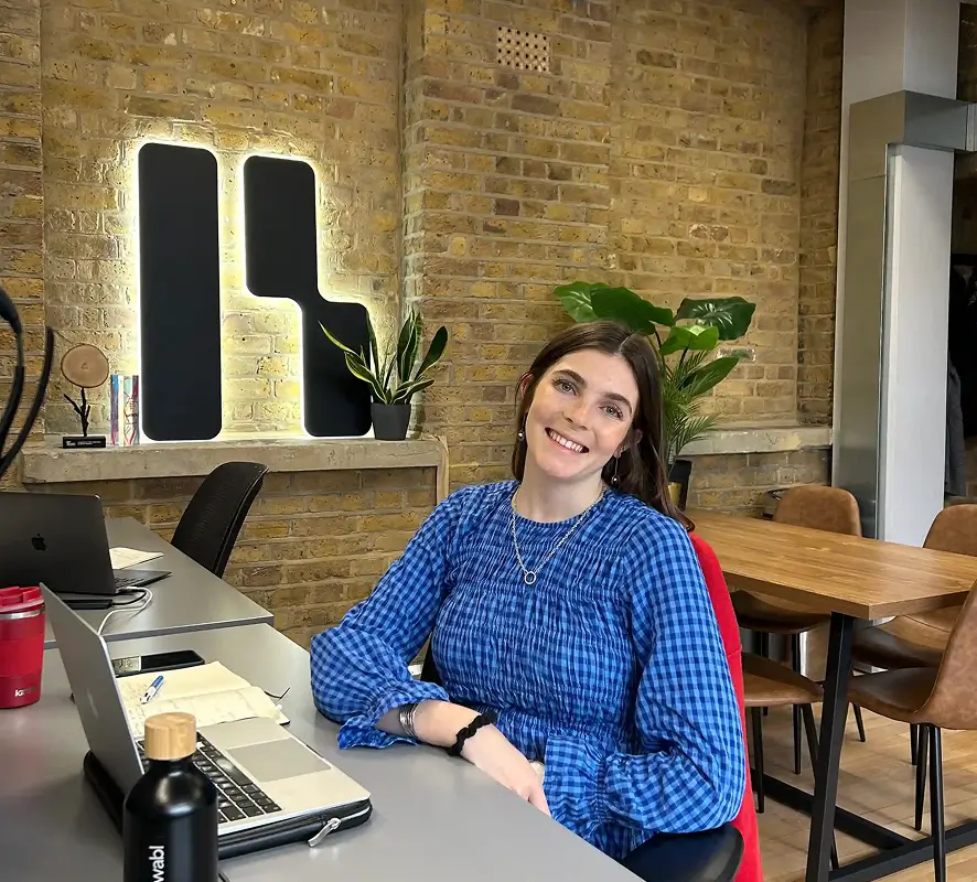 Renewable team photo | Woman in a blue checkered blouse sitting at a gray desk with a laptop and smiling in a modern office with exposed brick walls.