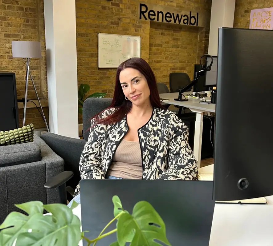 Renewable team photo | Woman with long dark hair sitting at a desk in an office, smiling at the camera with a laptop and monitor in front of her.