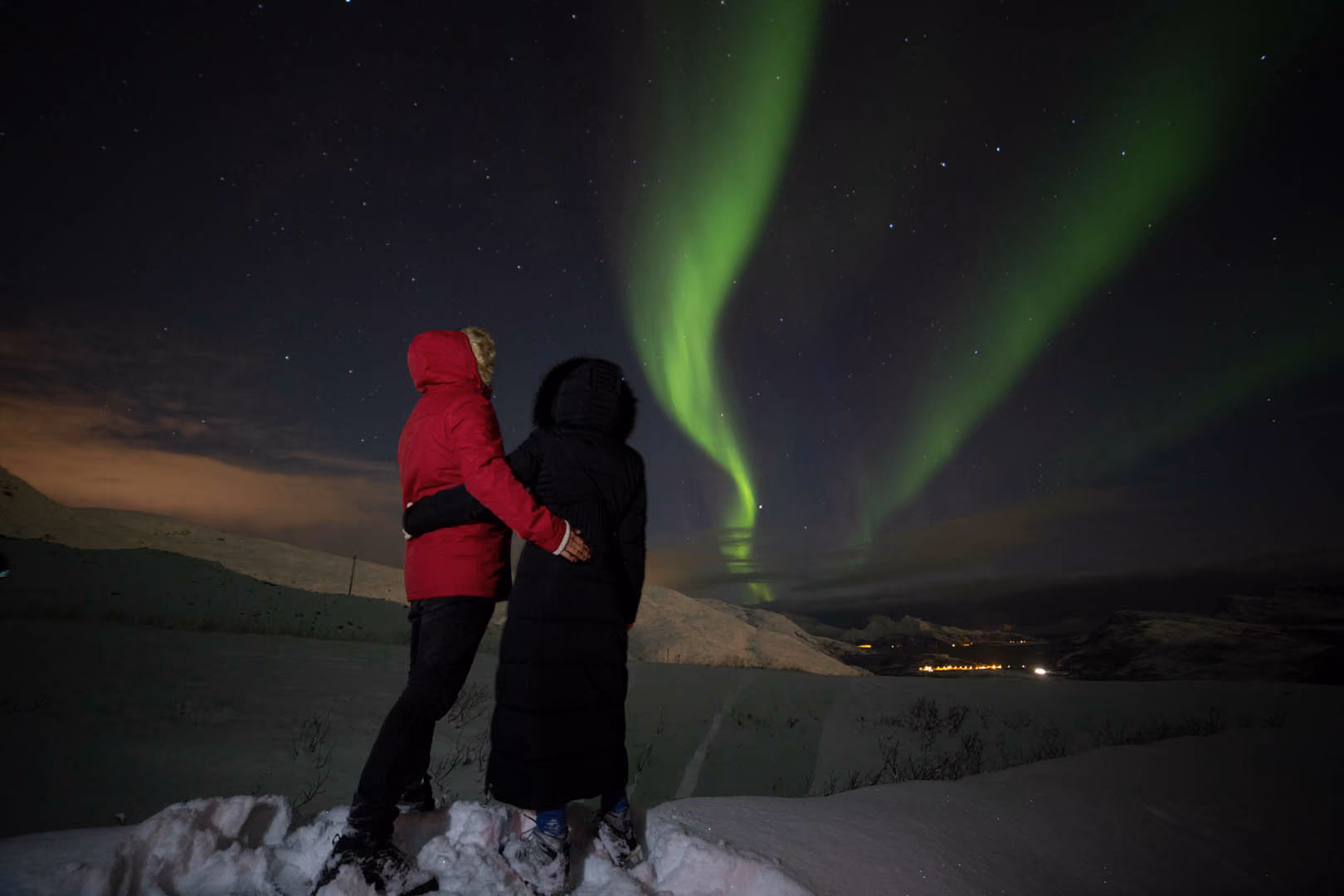 A couple dressed in winter coats stands on snow, embracing and watching the green northern lights in a starry night sky over snow-covered hills and distant village lights.