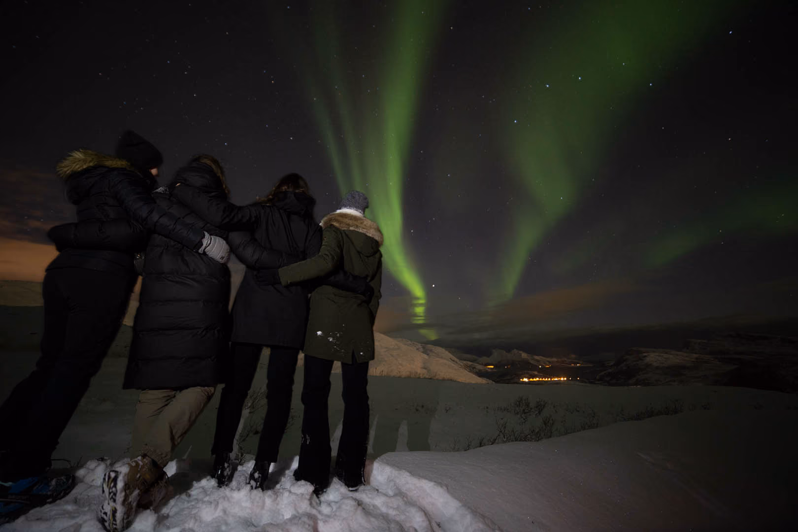 Four people wearing winter coats standing in snow with arms around each other, facing a night sky displaying bright green northern lights over snowy mountains and distant town lights.