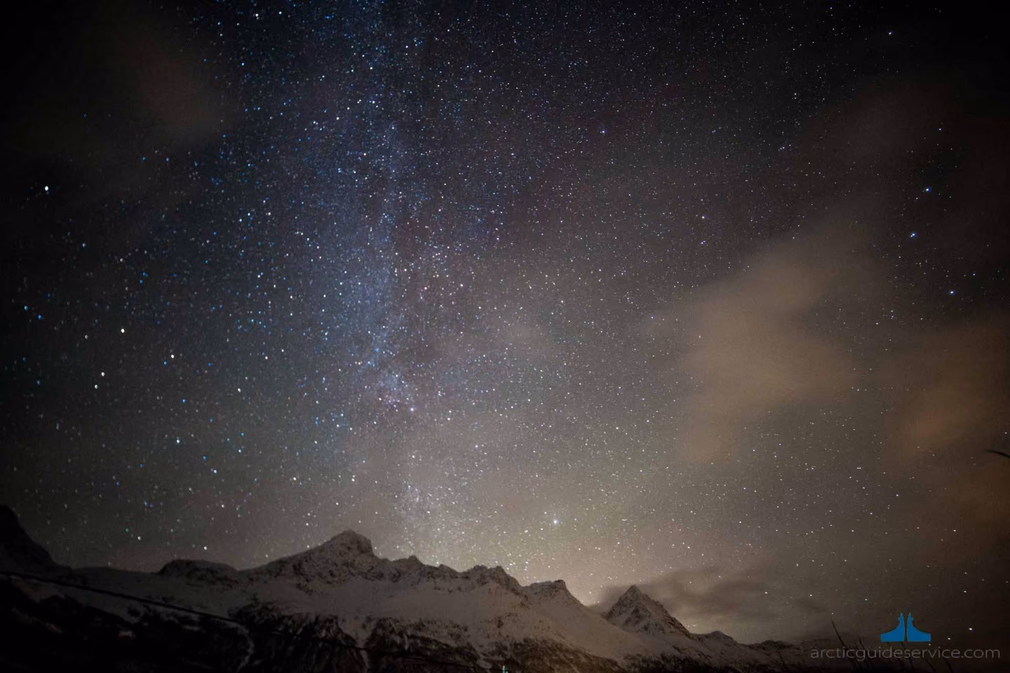 Snow-covered mountain peaks under a starry night sky with a faint Milky Way and some scattered clouds.