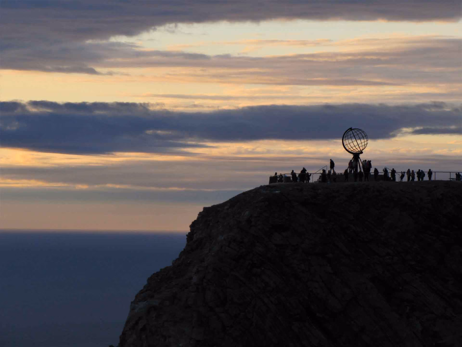 Silhouettes of people gathered around a globe sculpture on a cliff at sunset with a cloudy sky and ocean in the background.