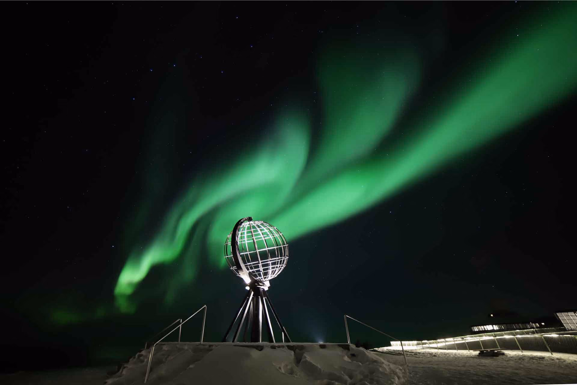 Illuminated metallic globe sculpture on a snowy hill under a night sky with bright green northern lights and stars, with a lit structure on the right.