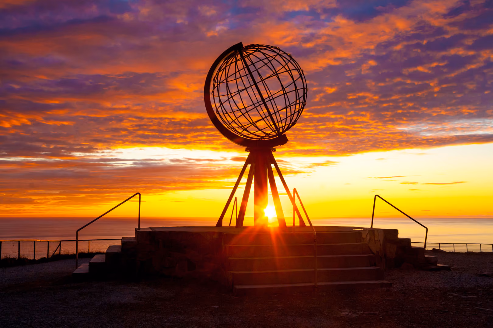 Sunset behind a large metal globe sculpture set on a raised platform with stairs and railings overlooking the sea.