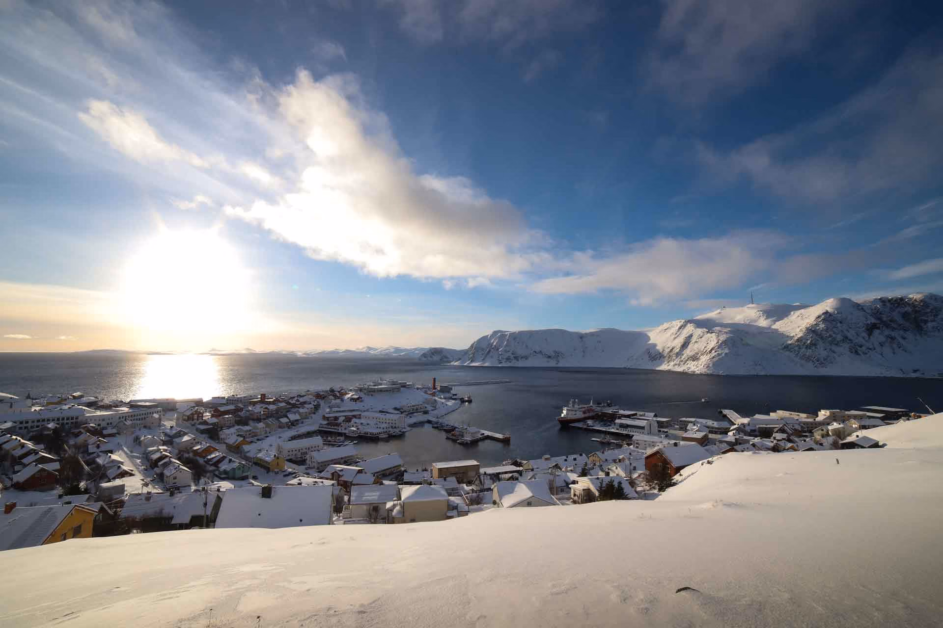 Snow-covered town by a calm body of water with ships docked at piers, set against snow-capped mountains under a partly cloudy blue sky with the sun low on the horizon.