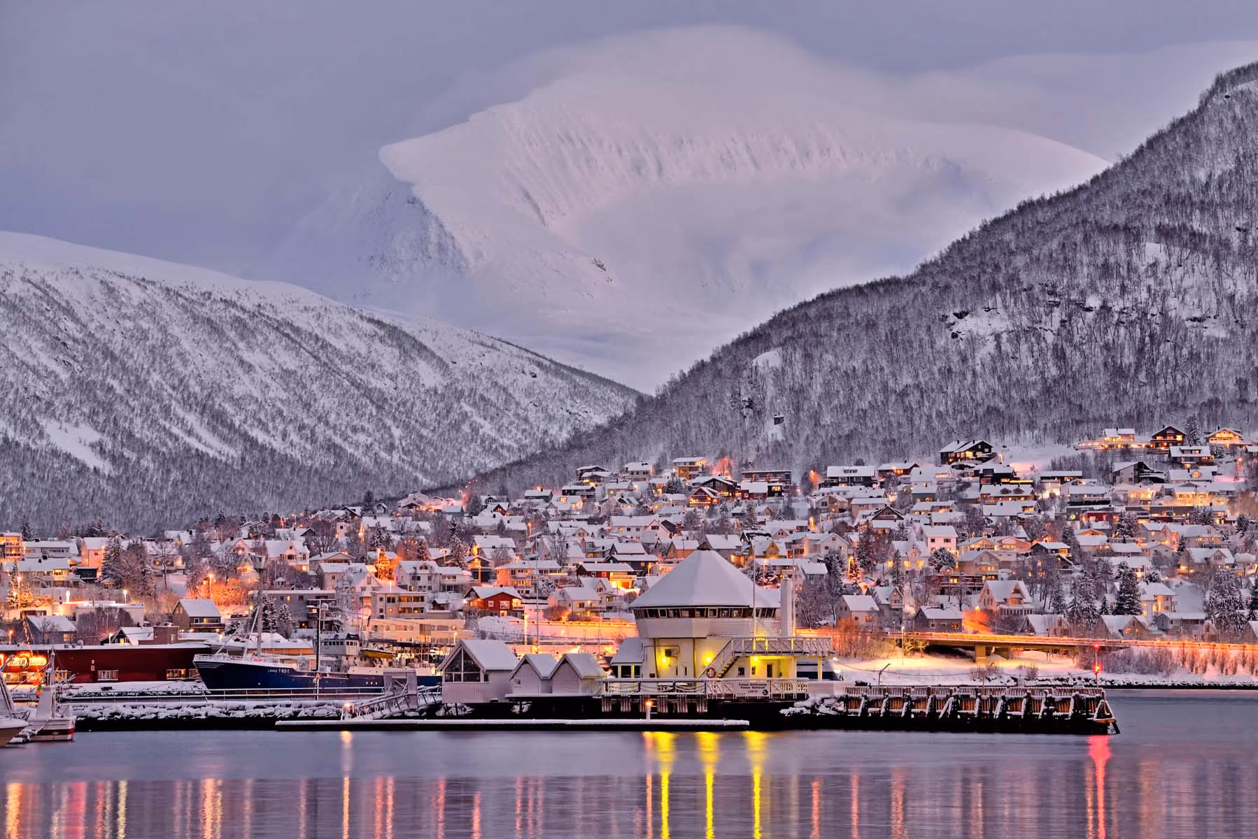 Snow-covered town with lit houses by water, surrounded by snowy mountains under a purple sky.