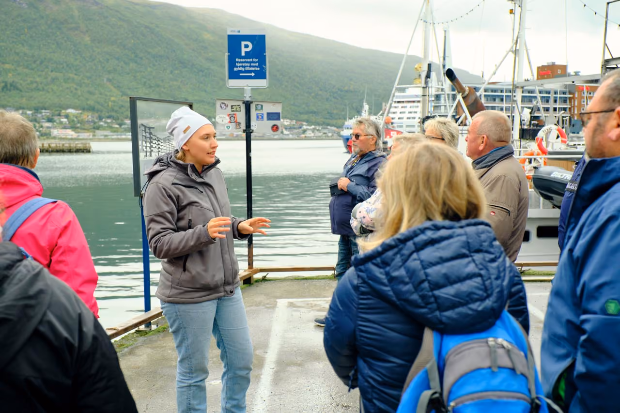 A woman in a gray jacket and white hat talks to a group of people standing by a waterfront with boats and a hillside in the background.