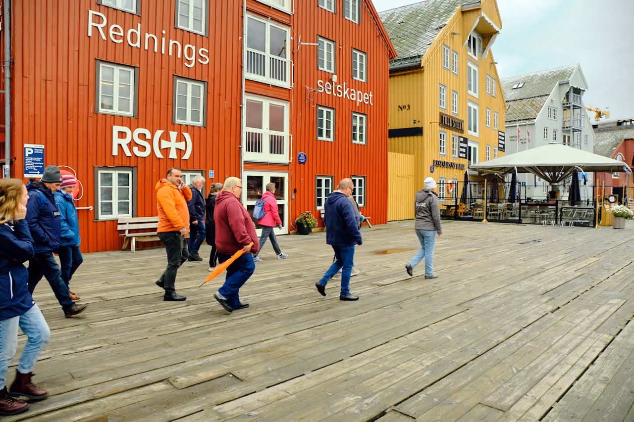 A group of people walking on a wooden boardwalk past colorful Scandinavian-style buildings painted red, yellow, and gray with signs including Rednings selskapet and Full Steam Restaurant & Bar.