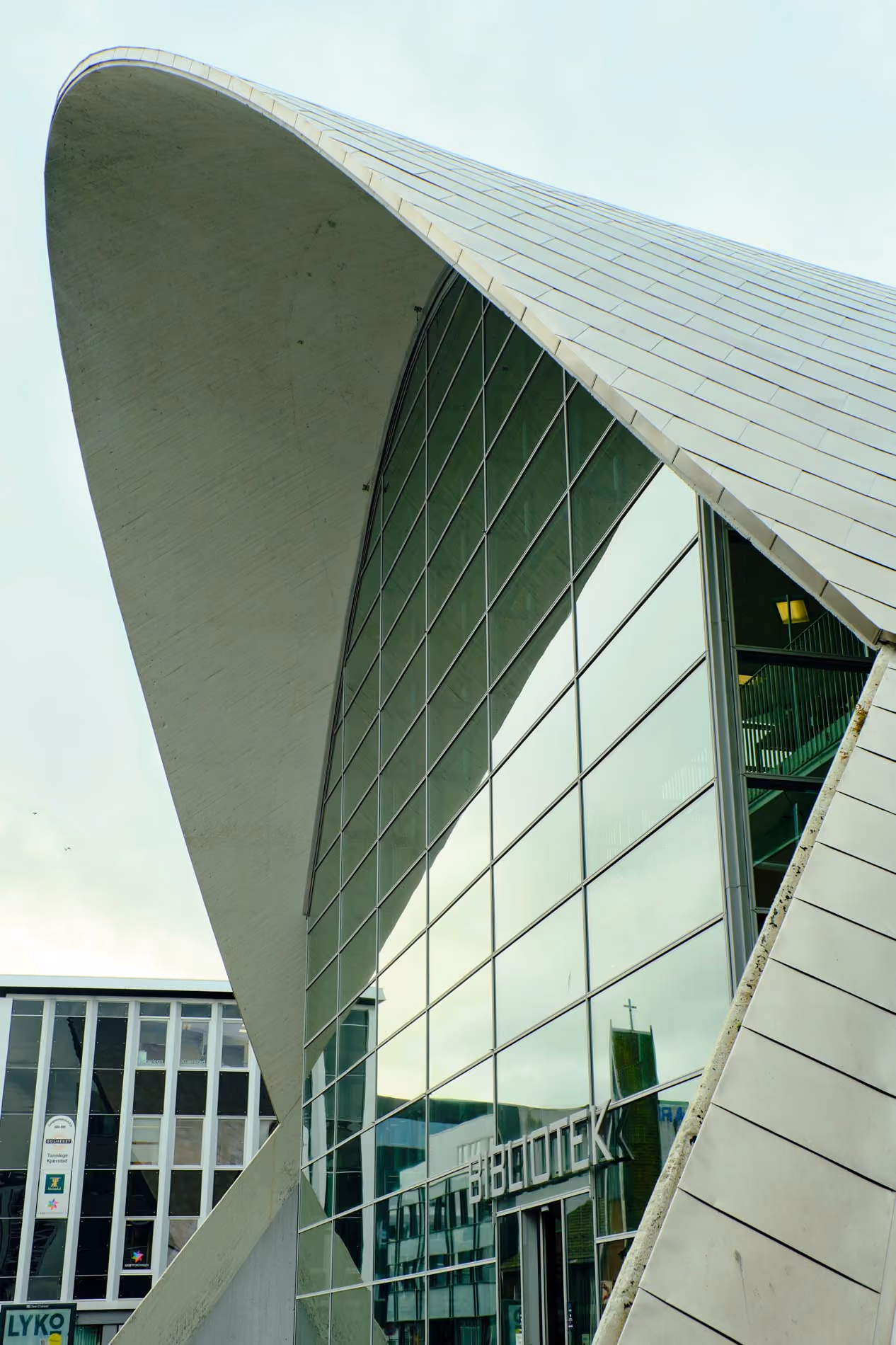 Modern building with a curved concrete roof and large glass windows reflecting nearby buildings, with the word 'BIBLIOTEK' visible on the glass entrance.