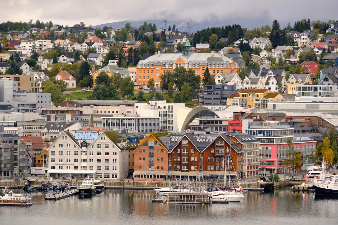 View of a waterfront city with boats docked by wooden piers, multi-story modern and traditional buildings, and a hillside of colorful houses and trees under a cloudy sky.