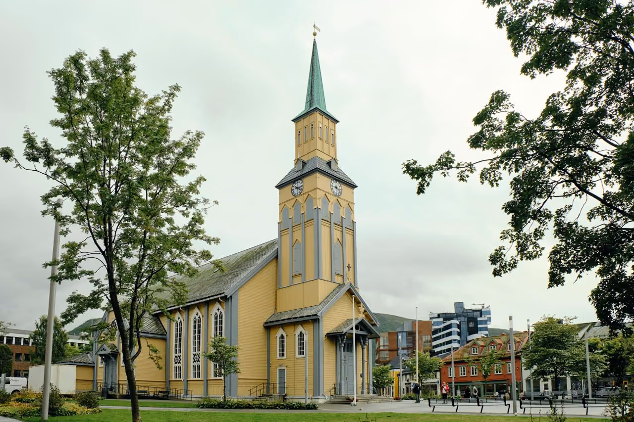 Yellow wooden church building with a tall greenish steeple and clock faces, surrounded by trees and benches in an urban park area with modern and traditional buildings in the background under an overcast sky.
