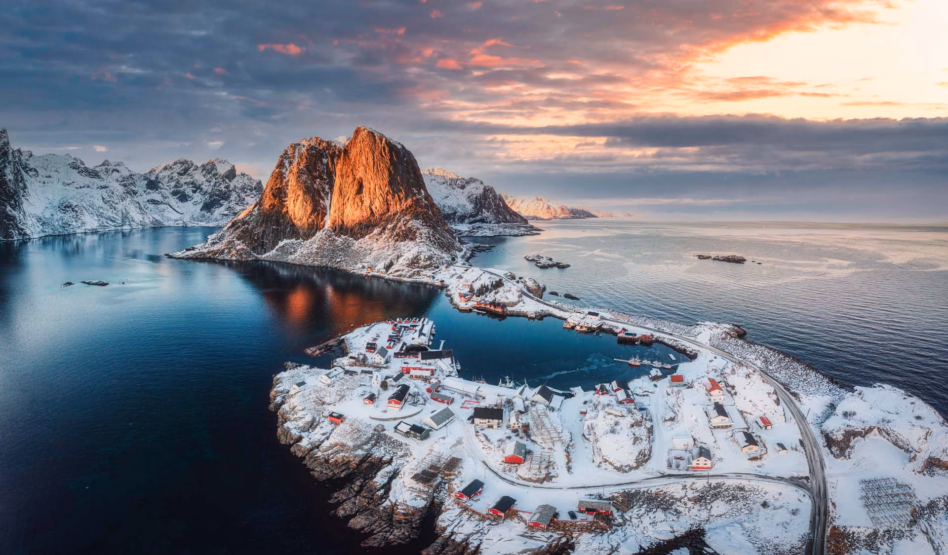 Snow-covered village by fjord with towering sunlit mountain and calm water under a cloudy sky.