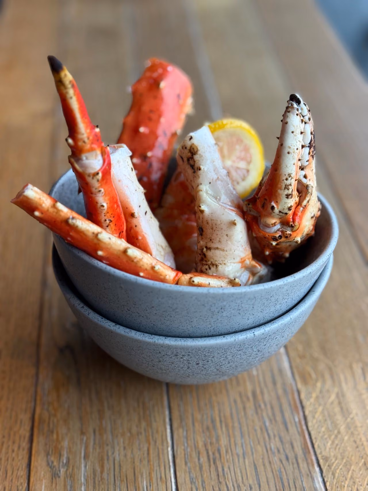 Steamed crab legs arranged upright in a gray bowl with a lemon slice, placed on a wooden table.