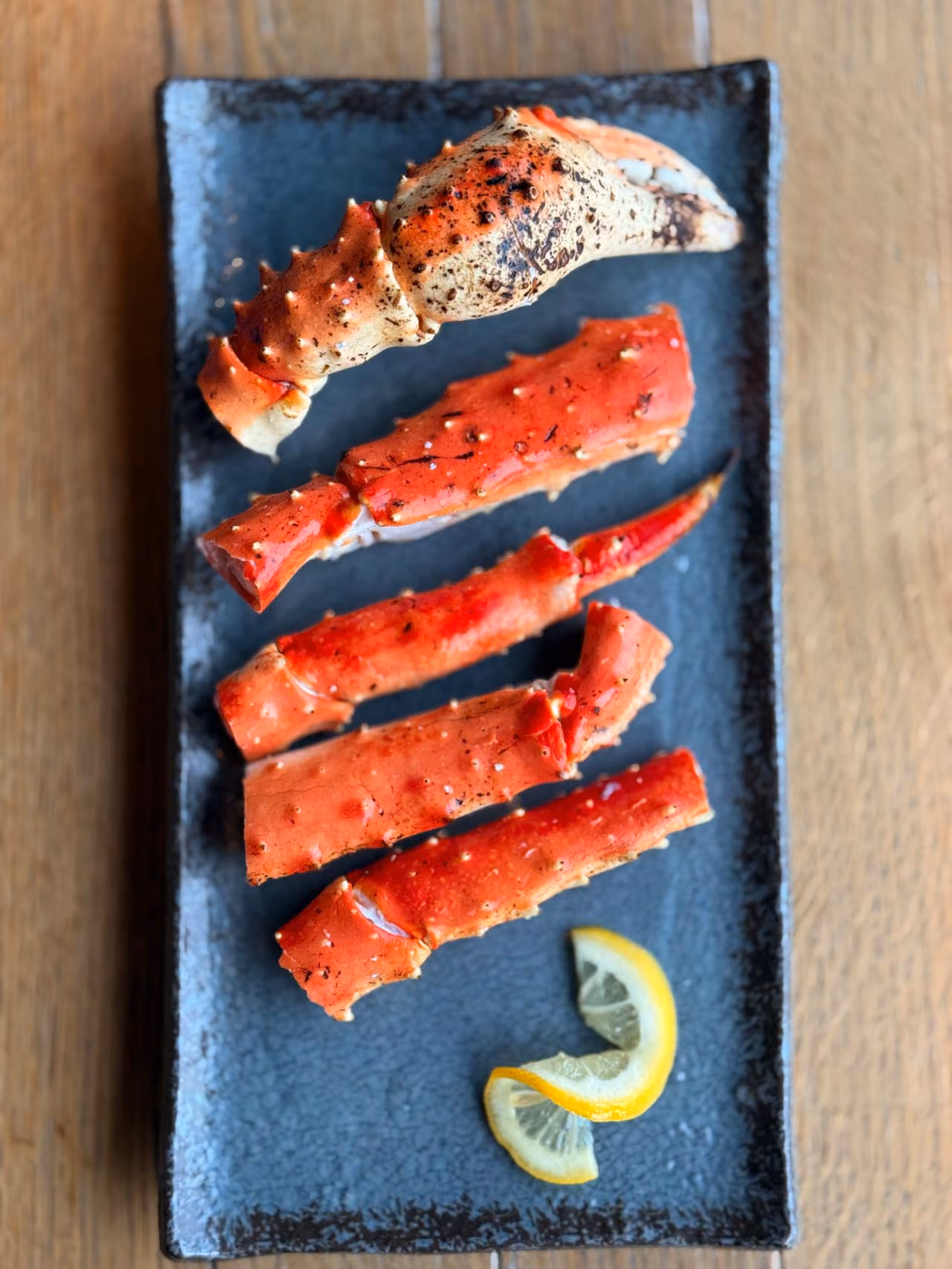 Cooked crab legs arranged on a dark rectangular plate with two lemon wedges on the side.