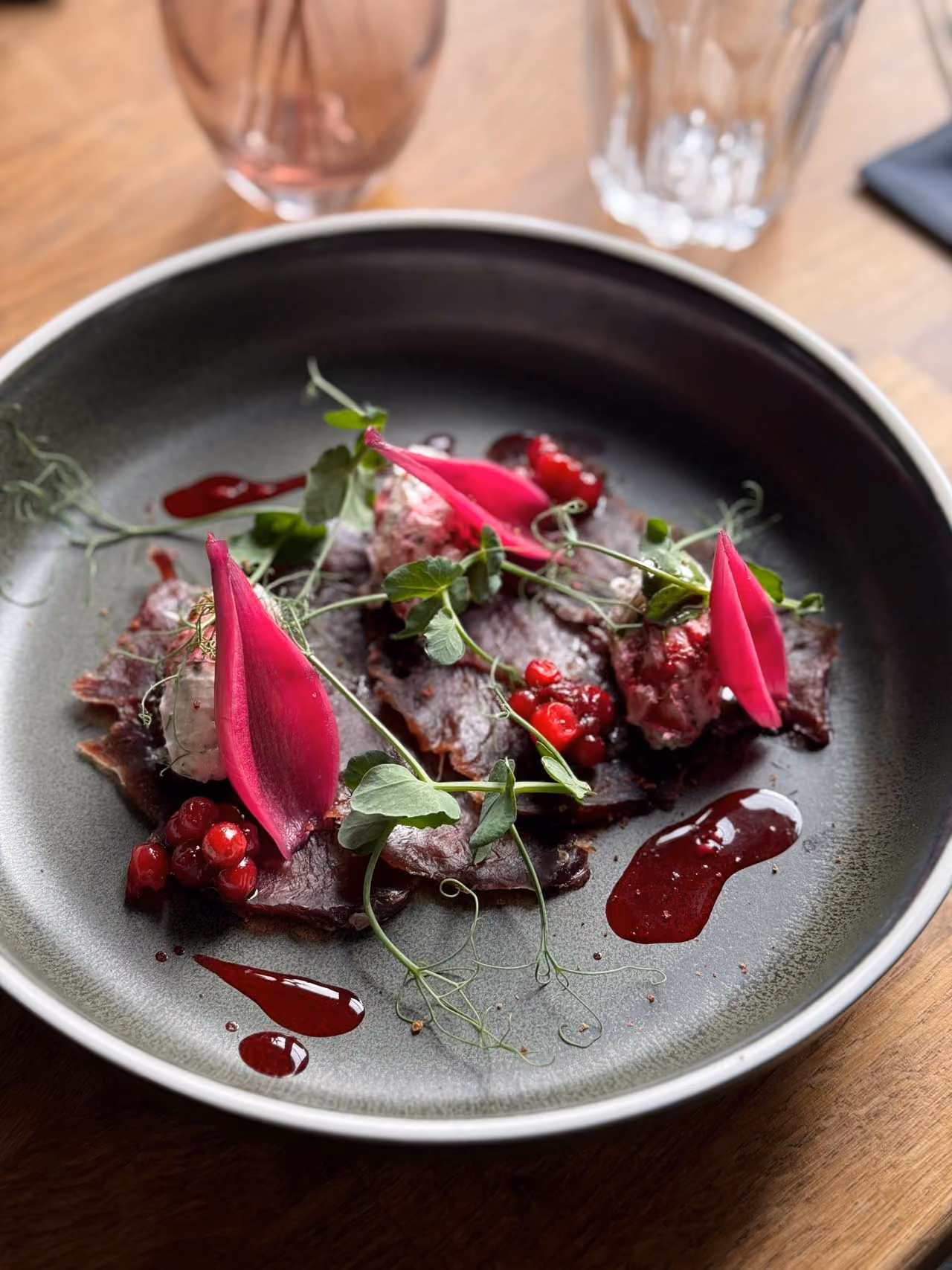 Plate with thin slices of dark meat topped with pink petals, berries, green herbs, and red sauce drops.