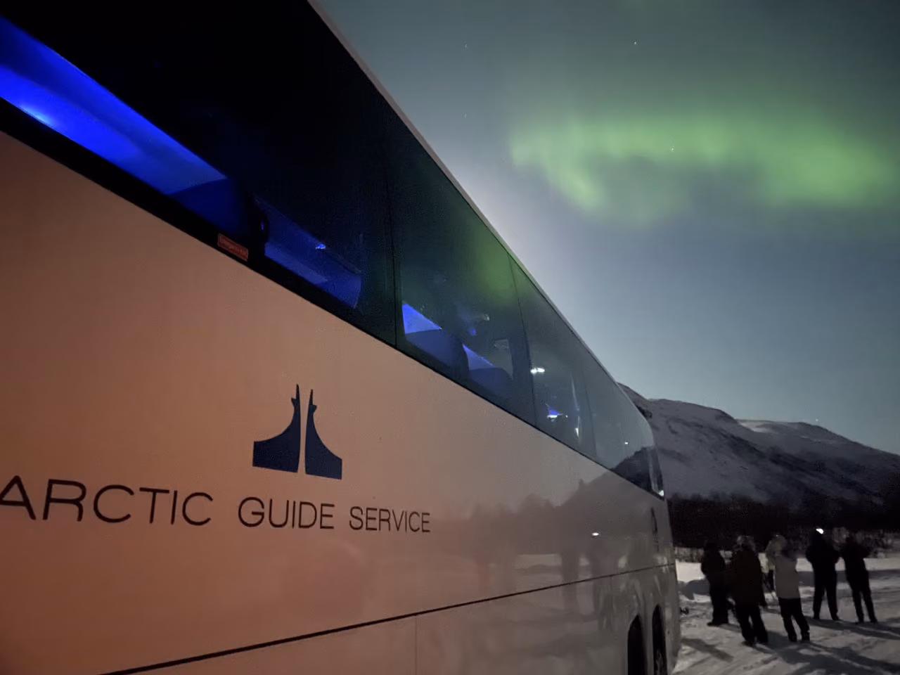 Bus with Arctic Guide Service logo parked at night under green northern lights with people nearby.