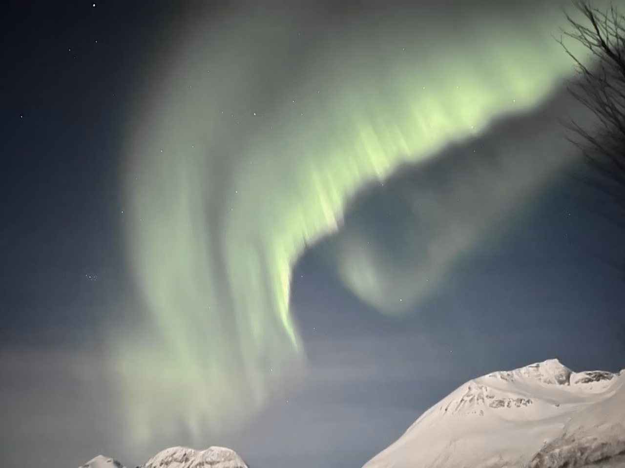 Green aurora borealis glowing over snow-covered mountain peaks at night with stars visible.