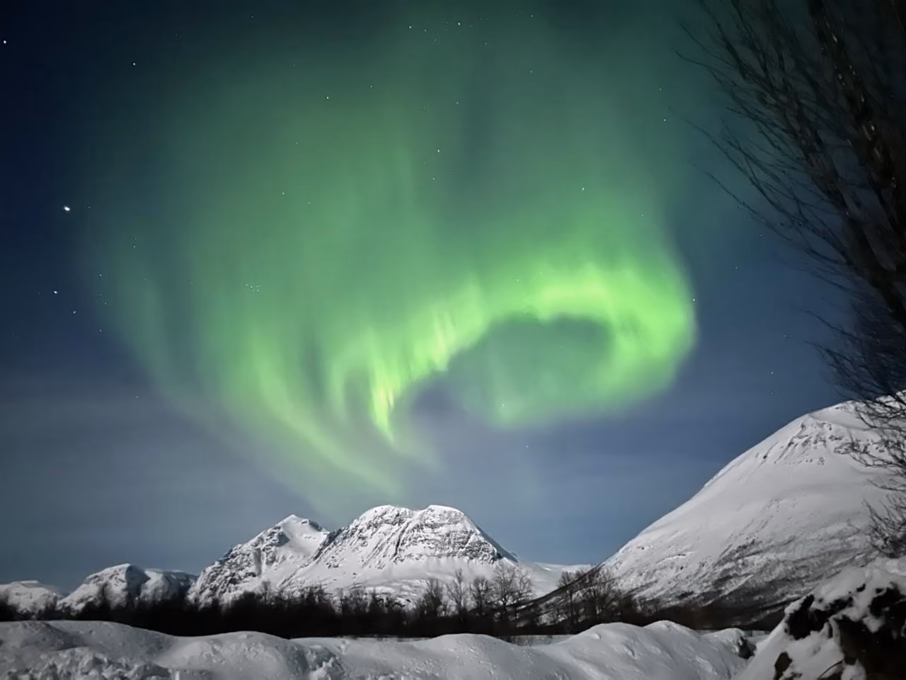 Green northern lights glowing and swirling above snow-covered mountains and a snowy landscape at night.