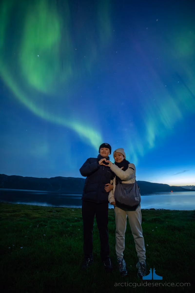 A couple standing on grass near a body of water at night making a heart shape with their hands, with the northern lights glowing green in the sky behind them.