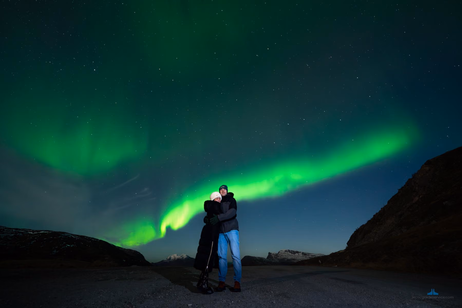 Couple embracing outdoors at night with a vivid green aurora borealis visible in the starry sky above snowy mountains.