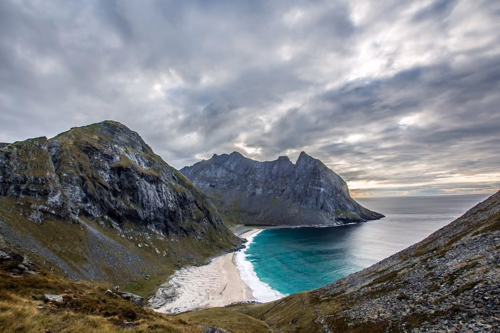 A serene curved sandy beach bordered by turquoise water and surrounded by steep rocky mountains under a cloudy sky.