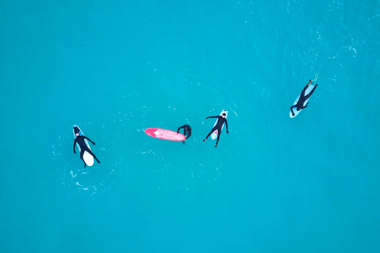Four surfers in wetsuits on surfboards floating in clear turquoise water seen from above.