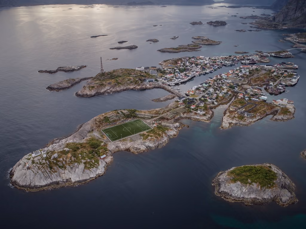 Aerial view of a small coastal village on rocky islands connected by bridges, with a green soccer field visible on one island surrounded by dark blue ocean water.