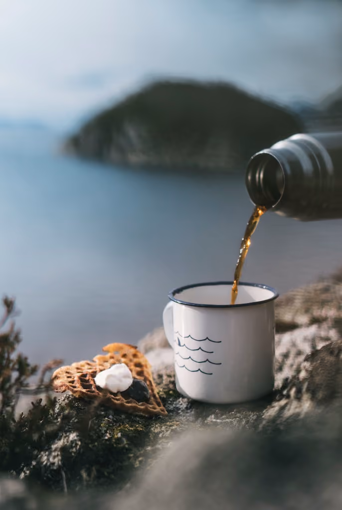 Pouring hot drink from flask into white enamel cup beside two heart-shaped waffles with cream by a lake.