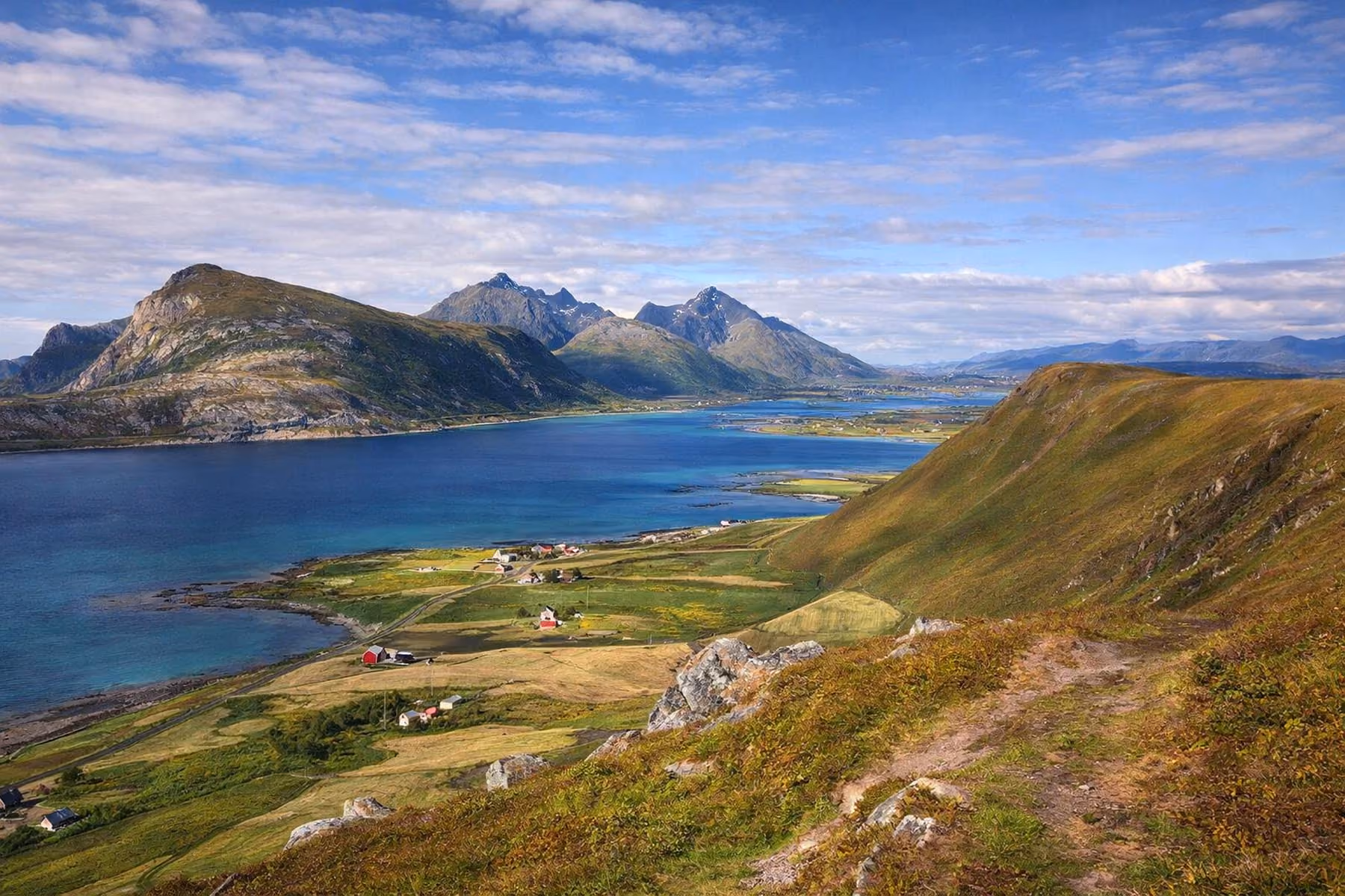 Scenic landscape view of a blue fjord surrounded by green hills and mountains with scattered houses under a partly cloudy sky.