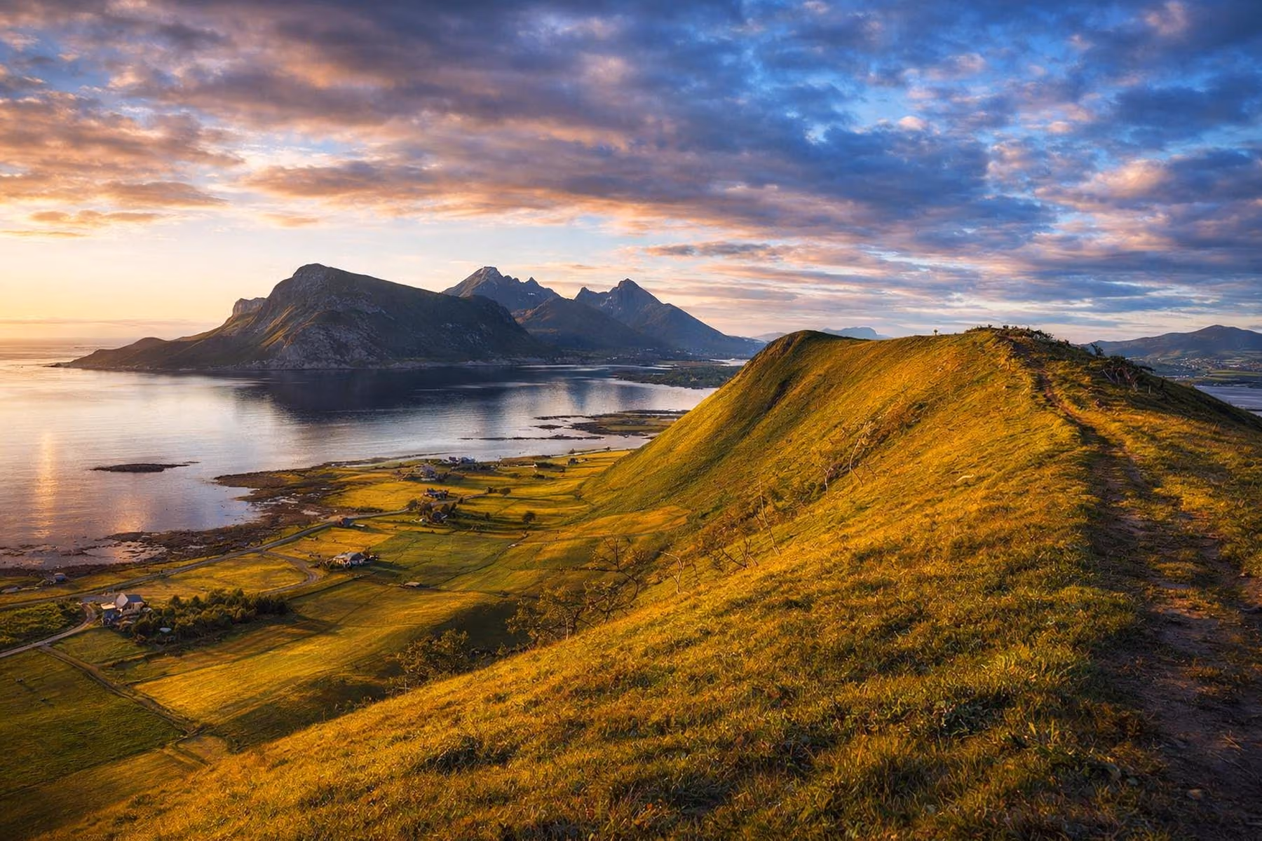 Sunlit grassy ridge overlooking a coastal valley with scattered houses and calm water, with mountains in the distance under a partly cloudy sky.