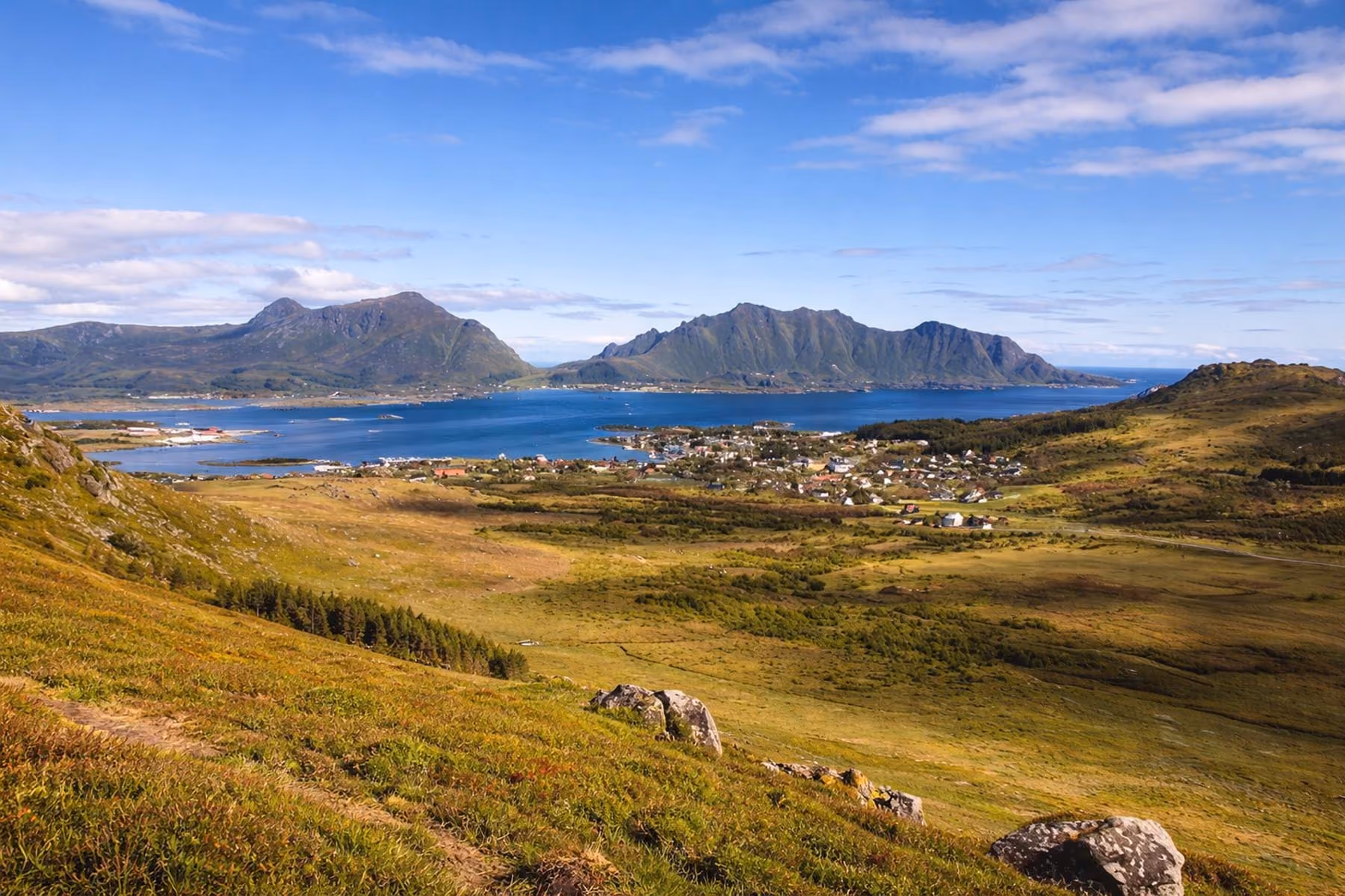 Panoramic view of a coastal village with clusters of houses near a calm blue bay, surrounded by grassy hills and rugged green mountains under a partly cloudy blue sky.