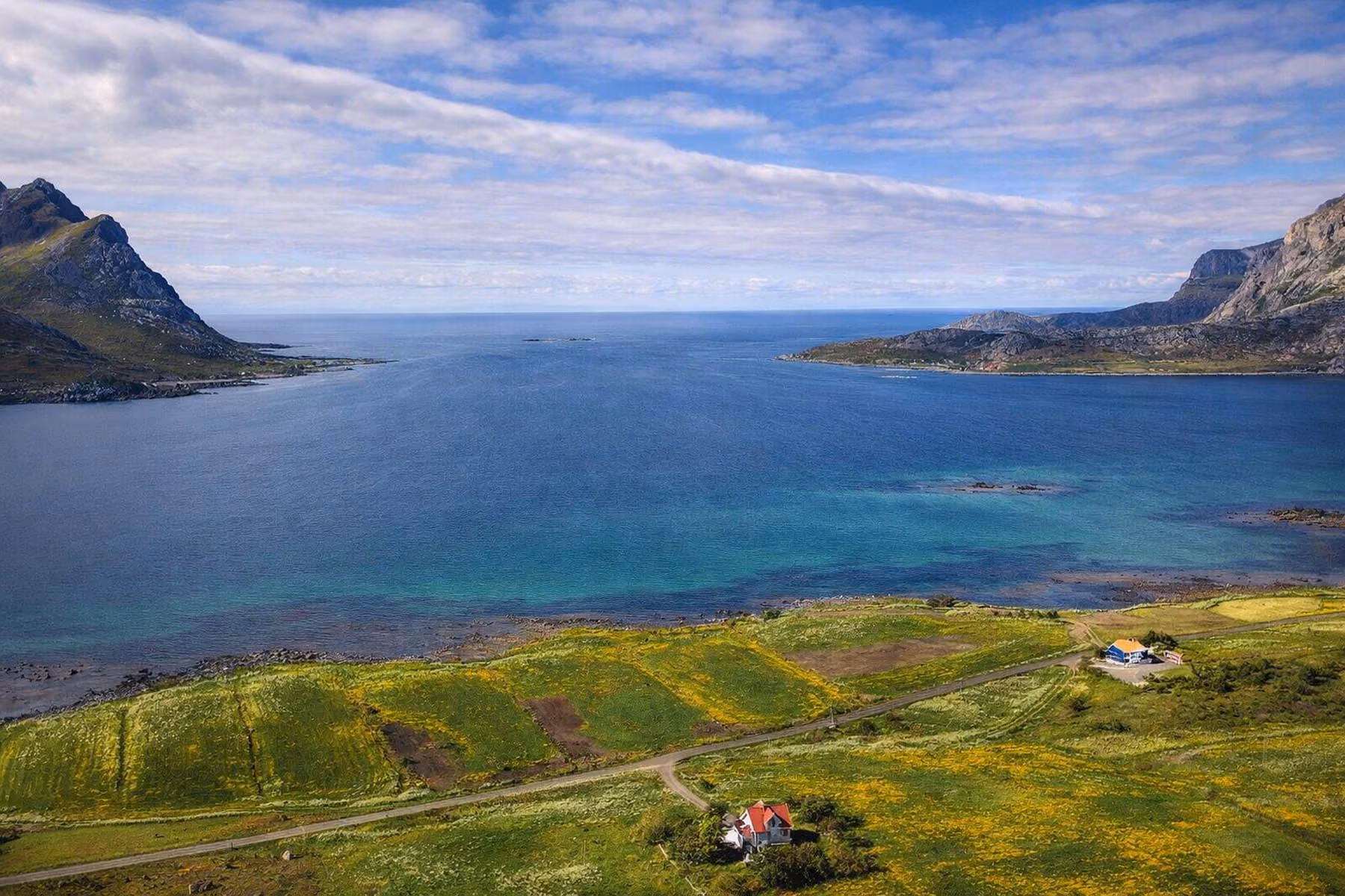 A coastal landscape with clear blue sea bordered by green fields and rocky hills under a partly cloudy sky, featuring two houses near the shoreline.
