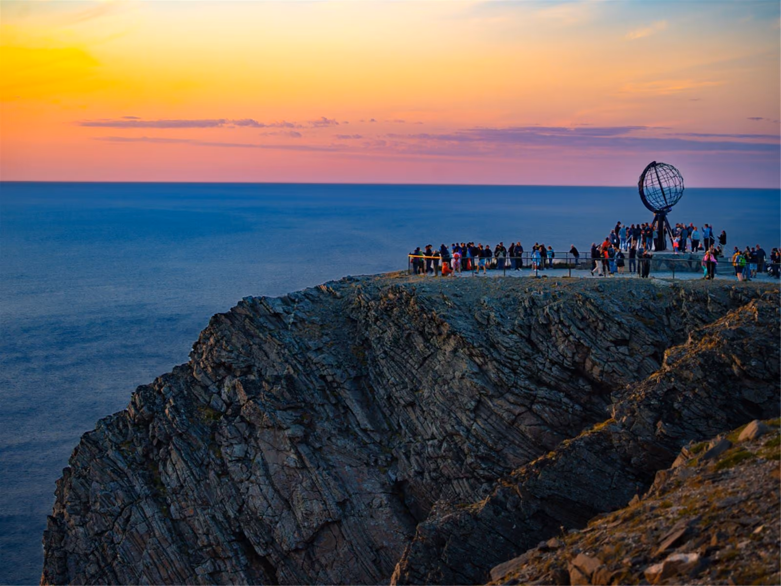 People gather near a globe sculpture on a rocky cliff at sunset by the ocean.
