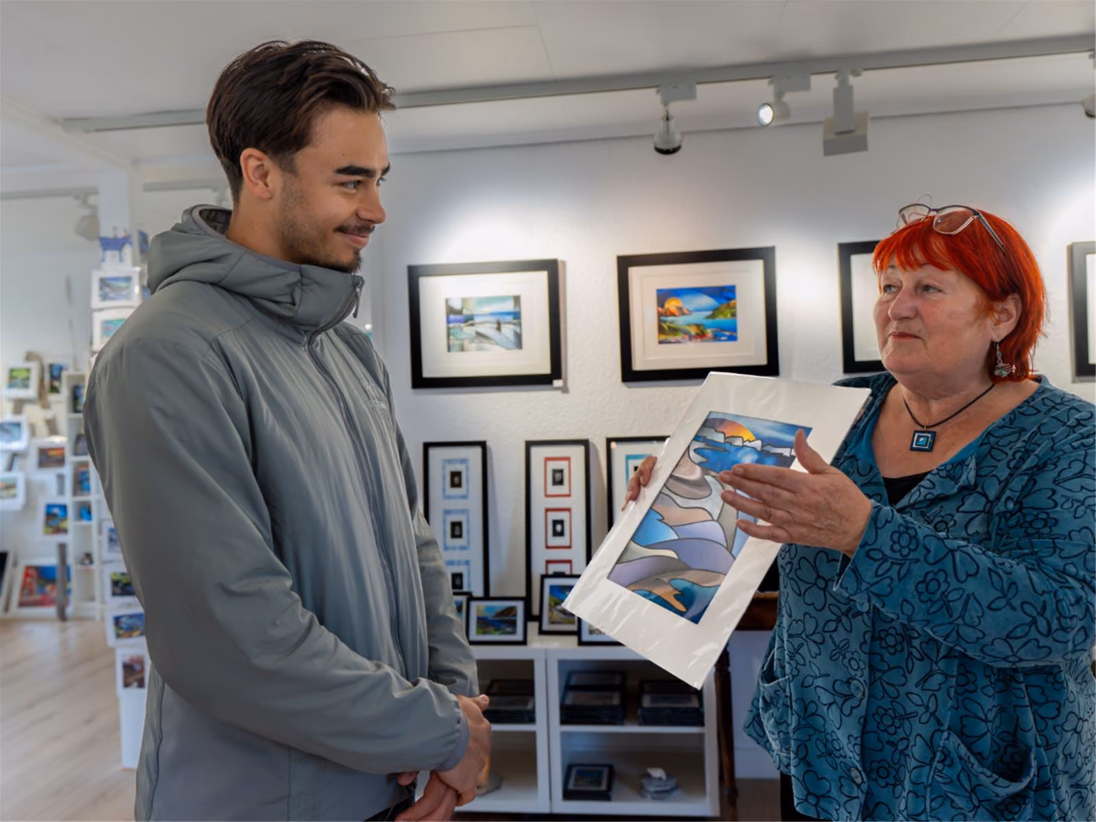 An older woman with red hair shows a colorful abstract landscape painting to a younger man in a gray jacket inside an art gallery with framed artwork on the walls.