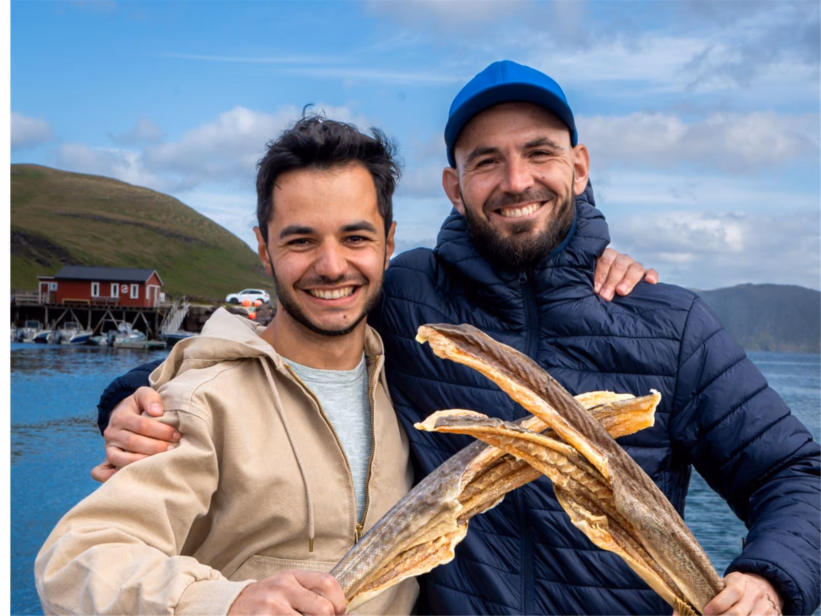 Two smiling men outdoors near water with boats, one wearing a blue cap and jacket holding dried fish, the other in a beige jacket with his arm around the other man's shoulder, hills and a red building are visible in the background.