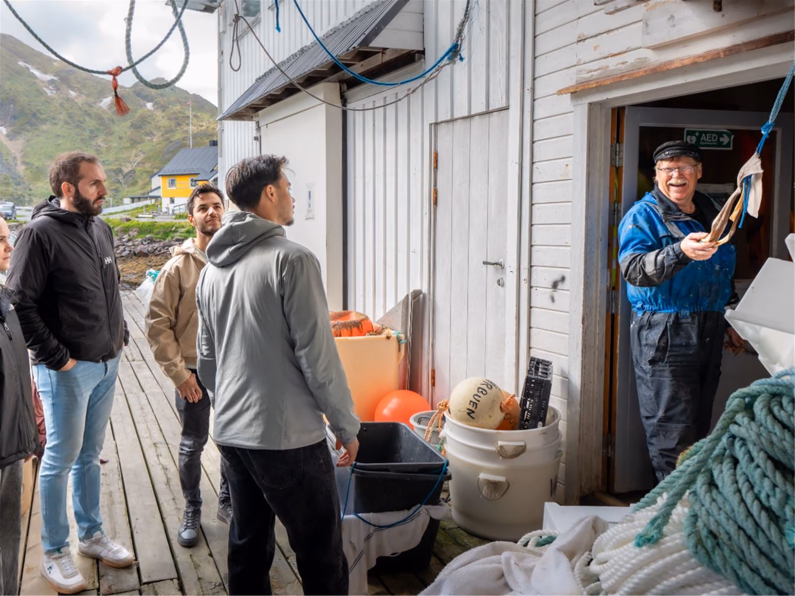 Group of people listening to a smiling fisherman holding equipment outside a wooden building.