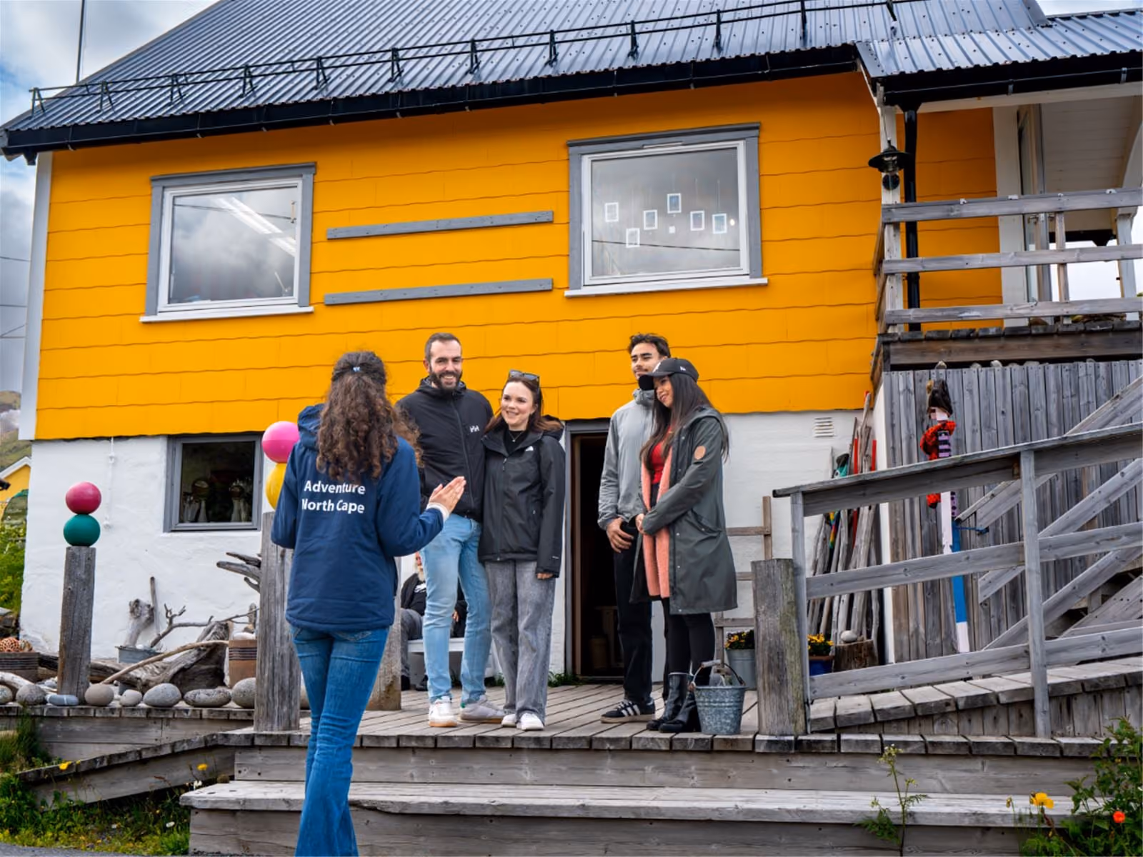 Four people standing on a wooden porch in front of a yellow and white house with a black roof, listening to a woman with curly hair wearing a blue jacket labeled 'Adventure North Cape' on the back.