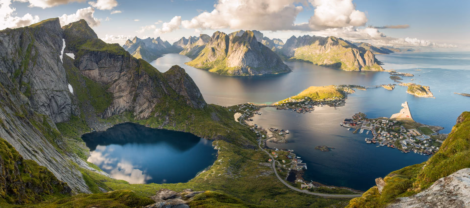 A panoramic coastal landscape with sharp mountains, a reflective lake, and a small village by the sea.