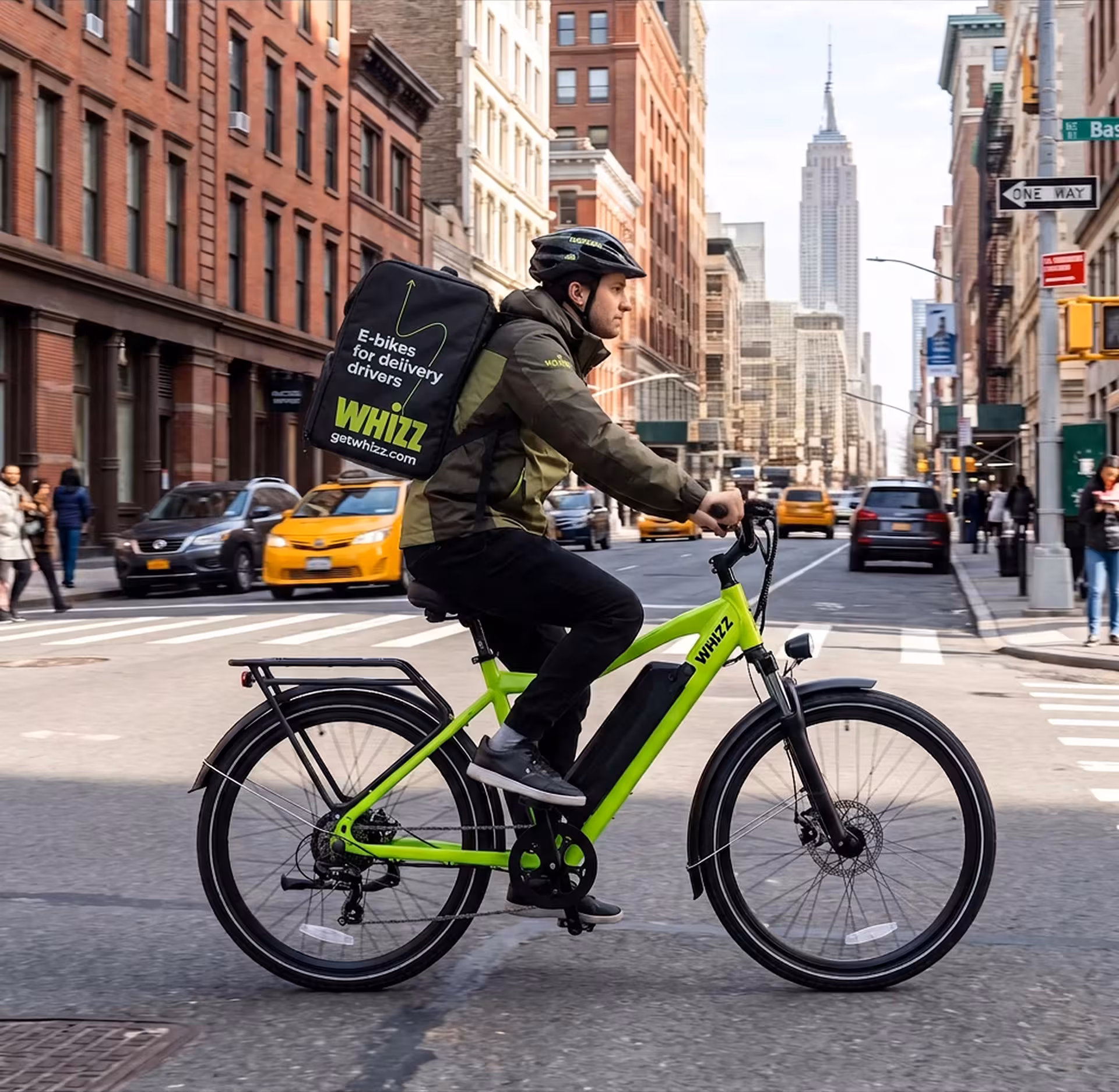 Side view of a delivery courier riding a neon-green electric bicycle on a city road. The rider wears a helmet and a black backpack with text "E-bikes for delivery drivers WHIZZ". Classic NYC brick buildings and the Empire State Building are visible in the background.