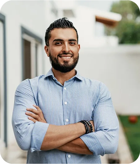 A person with crossed arms stands confidently outdoors, wearing a button-down shirt with vertical stripes in a serene setting.