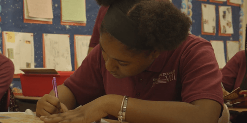 Student writing at desk in classroom with bulletin board behind her