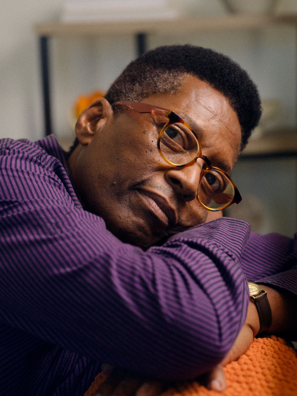 A man wearing a purple shirt sitting on a couch and resting his head on his forearms 